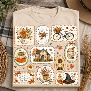May include: A beige t-shirt featuring a collage of autumn-themed illustrations in scalloped frames, including a house, a ghost, a bicycle with a pumpkin basket, a mug of cocoa, a lantern, a witch hat, and flowers with the text Crisp leaves Warm hearts Cozy days.