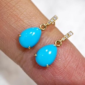 May include: A pair of gold drop earrings featuring smooth, teardrop-shaped turquoise stones suspended from thin, vertical bars accented with small, clear, sparkling stones. The earrings have a polished gold finish and a delicate, refined structure.