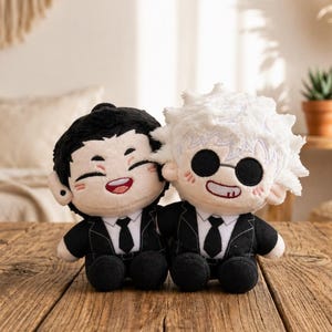 Puede incluir: Dos muñecos de peluche de personajes de anime, uno con pelo negro y otro con pelo blanco y gafas de sol. Ambos muñecos visten trajes negros y corbatas, con caras sonrientes. Están sobre una superficie de madera.