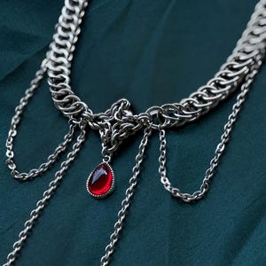 May include: A silver-toned necklace with a chainmail-style collar and dangling chains. A teardrop-shaped, red gemstone pendant hangs from the center. The necklace is displayed against a dark teal fabric background.