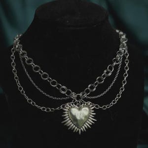 May include: A multi-layered silver-toned chain necklace featuring a central heart-shaped pendant with radiating spikes. The design consists of a thick circular link chain and thinner delicate chains that drape to support the metallic heart charm.