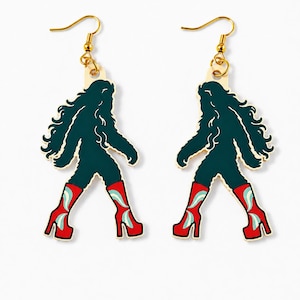 Pode incluir: Um par de brincos pendentes com uma figura humanoide peluda verde-escura usando botas de plataforma vermelho-brilhante com detalhes em azul-claro. Os brincos estão suspensos em ganchos de metal dourado e têm uma base de madeira clara.