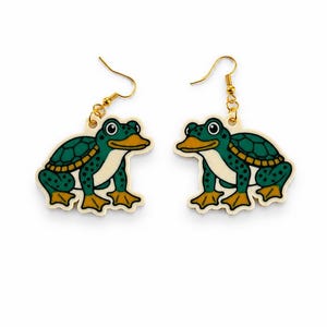 May include: A pair of whimsical frog-turtle hybrid earrings. Each earring features a cartoon frog with a turtle shell design, in shades of green, yellow, and white. The earrings have gold-colored fish hook closures.