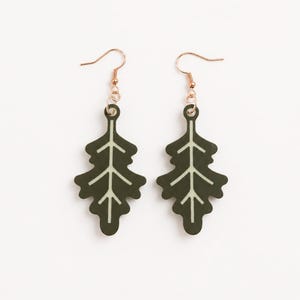 May include: A pair of dangle earrings featuring dark olive green oak leaf shapes with a light cream-colored central vein detail. The leaf pendants are suspended from rose gold-toned metal fishhook wires, creating a symmetrical set of earrings.