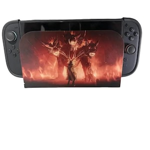 May include: Black Nintendo Switch console with a custom cover featuring a fiery scene with a three-headed creature and a figure. The cover is inserted into the console's central slot, enhancing its appearance.