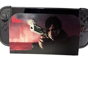 May include: A black Nintendo Switch console with a screen protector featuring a man holding a gun. The screen protector has a red and black background. The console has two gray controllers attached.