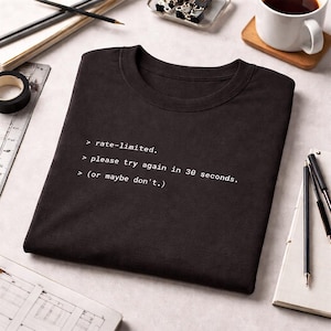 May include: Black t-shirt with white text: "> rate-limited. > please try again in 30 seconds. > (or maybe don't.)". The shirt is folded on a light surface, with office supplies and a coffee cup in the background.