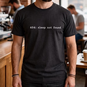 May include: Black t-shirt with the text "404: sleep not found" in white. The shirt is worn in a coffee shop setting. Other people are visible in the background, along with coffee cups and a laptop.