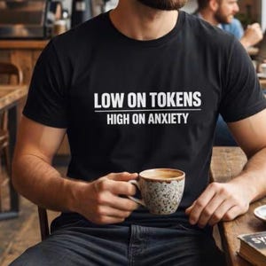 Puede incluir: Camiseta negra con el texto "LOW ON TOKENS HIGH ON ANXIETY" en blanco. La persona sostiene una taza de café moteada. La escena está ambientada en una cafetería con mesas y sillas de madera. Un libro titulado "SYNTHETIC DREAMS" está sobre la mesa.