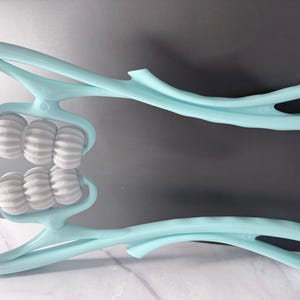 May include: A light blue facial massager with two sets of white rollers. The massager has a curved handle and is designed to be held in the hand. The rollers are ribbed and appear to be made of a hard plastic material.