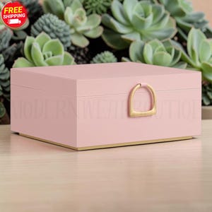 May include: A rectangular light pink storage box with a smooth finish, featuring a gold-toned stirrup-shaped latch on the front and a gold-toned base. The box has a clean, structured design with a flat lid and a solid, uniform pastel color.