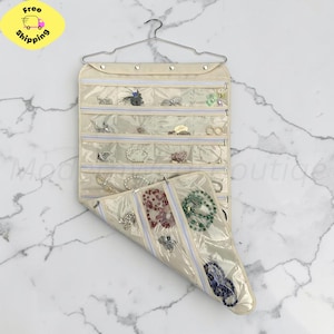 May include: A beige hanging jewelry organizer with multiple clear plastic pockets and zippered compartments. The organizer is suspended from a metal hanger and contains various pieces of jewelry, including necklaces, bracelets, and rings.