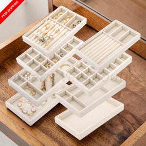 May include: A modular jewelry organizer set featuring multiple stackable trays in a light beige finish. The trays include various compartments, including ring rolls, square grids, and open rectangular sections, containing assorted gold-toned jewelry.