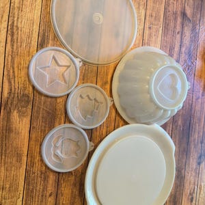 Puede incluir: Colección de utensilios de cocina de plástico blanco roto vintage, que incluyen tapas, un molde en forma de estrella, un molde en forma de corazón y una fuente ovalada. Los artículos están dispuestos sobre una superficie de madera.