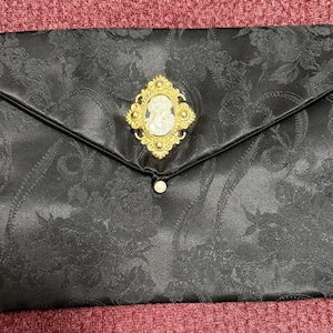 May include: Black satin clutch with a floral pattern and a gold-toned cameo brooch. The brooch features a white cameo portrait within a gold frame. The clutch has a flap closure with a small pearl-like button.