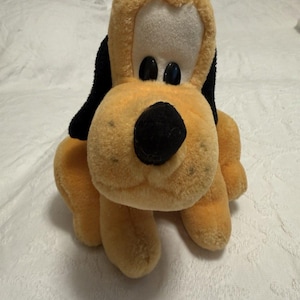 May include: A plush toy dog with a tan body, black ears, and a black nose. The dog has large, black eyes and a white face. The toy is sitting on a white surface.