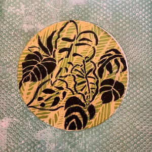 May include: A round, botanical-themed design featuring black leaf illustrations on a yellow-green background. The design is set against a textured, light green backdrop. The artwork has a hand-drawn aesthetic.