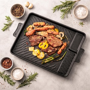 Può includere: Una piastra rettangolare nera con una superficie di cottura a coste, piena di carni grigliate, verdure e salsicce. Piccole ciotole di spezie, sale e rametti di rosmarino circondano la piastra.