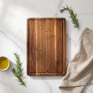 Puede incluir: Una tabla de cortar rectangular de madera con ranura para jugos, rodeada de ramitas de romero, un pequeño cuenco con aceite de oliva y un paño de lino beige. La tabla es de madera marrón oscuro y está sobre una superficie de mármol blanco.