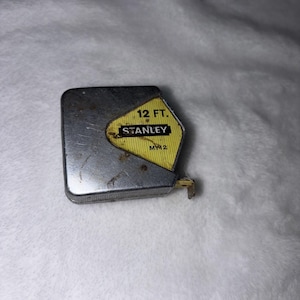 May include: A vintage Stanley 12 ft. (3.66 m) tape measure. The metal casing is silver with a yellow and black label. The label displays the Stanley brand name and measurement information. The tape measure is designed for measuring distances.