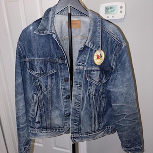 May include: A blue denim jacket with a button-up front, two chest pockets, and two side pockets. The jacket has a faded appearance and a small patch on the front. A Levi's brand tag is visible.
