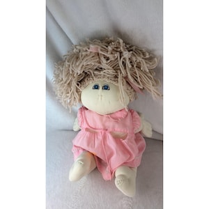 May include: A soft fabric doll with a round, pale face, blue eyes, and a small nose. The doll has thick, light beige yarn hair styled in a messy, textured mop. It wears a light pink, short-sleeved dress with a simple collar and gathered fabric.