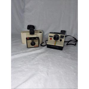May include: Three vintage Polaroid cameras are displayed against a white backdrop. One is a small, black and silver camera. Another is a cream-colored camera with a black strap and a rainbow stripe. A flash unit is also present.