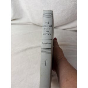 May include: A light gray hardcover book spine titled The Greatest Faith Ever Known by Fulton Oursler. The spine features black text, horizontal line accents at the top and bottom, and a small black cross symbol near the bottom edge of the book.
