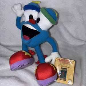 May include: A blue plush toy character with white, red, and yellow accents. The toy has two white heads with black eyes, a large open mouth, and red shoes. A tag is included.
