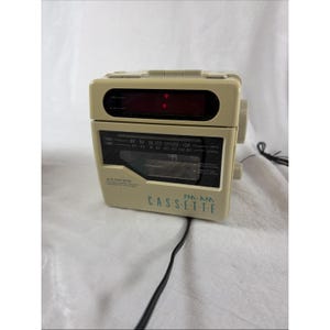 May include: A beige Lloyd's FM-AM cassette player with a black radio frequency display panel, a digital clock window at the top, and a black power cord extending from the bottom. The front features the text LLOYD'S FM-AM CASSETTE.