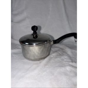 May include: A stainless steel saucepan with a lid and a black handle. The lid has a black knob. The saucepan is sitting on a white, textured surface. The handle has a loop at the end.