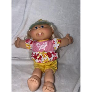 May include: A Cabbage Patch Kids doll with blonde hair and blue eyes. The doll wears a pink top with a butterfly graphic and yellow shorts. The top has a floral pattern with red and yellow flowers.