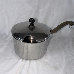 May include: A stainless steel saucepan with a lid and a long handle. The lid has a black knob and a small opening. The handle is brown and has a loop at the end. The saucepan is sitting on a white surface.