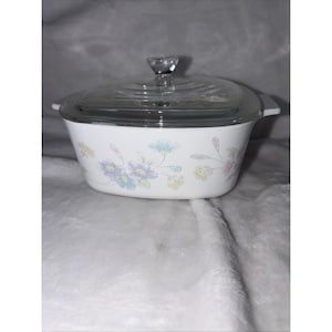 May include: White casserole dish with a clear glass lid. The dish is decorated with a floral pattern in pastel blue, purple, and yellow. The lid has a clear, diamond-shaped handle. The dish is oval-shaped and has two white handles.