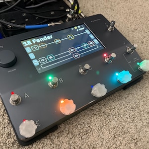 May include: A black guitar effects processor with a screen displaying a diagram. It has several knobs and foot switches with colored lights. The screen displays "1E Fender". The word "VOLUME" is printed on the device.