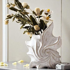 Könnte beinhalten: Eine weiße Keramikvase in Form einer spiralförmigen Muschel mit braunen, strukturierten Akzenten. Die Vase enthält einen Strauß aus getrocknetem grünem Laub, runden beigen Samenständen und weißen Blüten mit gelben Mitten.