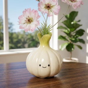 Puede incluir: Un jarrón de cerámica caprichoso con forma de cebolla, con una cara sonriente, que contiene flores rosas y blancas. El jarrón blanquecino tiene una parte superior verde y está sobre una mesa de madera. Una planta de interior está en el fondo.