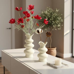 May include: Two cream-coloured, wavy-shaped vases with red flowers and cotton stems. A white candle sits on a marble tray. The vases are on a white table, with a potted plant in the background. The theme is home decor.