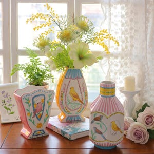 May include: Three decorative ceramic vases featuring bird designs. The vases are painted in pink, yellow, and orange, with blue and green details. One vase is rectangular, the others are rounded. Flowers and a candle are also present.
