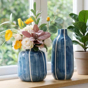 May include: Two blue ceramic vases with white vertical stripes. One vase contains a floral arrangement of yellow, white, and pink flowers. The vases are on a wooden surface near a window.