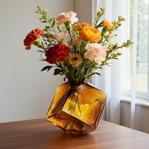 Peut inclure: Un bouquet de fleurs colorées dans un vase géométrique en verre ambré. Les fleurs comprennent des œillets orange, rouges et roses, et des fleurs jaunes et blanches. Le vase est angulaire et posé sur une surface en bois.