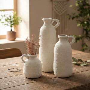 May include: Three off-white textured ceramic vases of varying heights with handles. The smallest vase holds dried pink stems. The vases are arranged on a wooden table, with a bracelet and greenery visible.