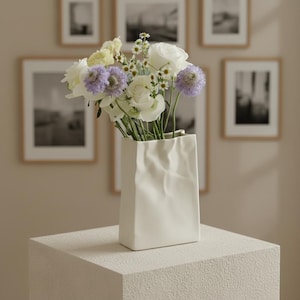 以下が含まれることがあります： 白のセラミック花瓶。くしゃくしゃの紙袋のデザインで、白いバラ、紫のオニグルミ、小さな白と黄色の花の花束が入っています。花瓶は、質感のある正方形の台座の上に置かれています。