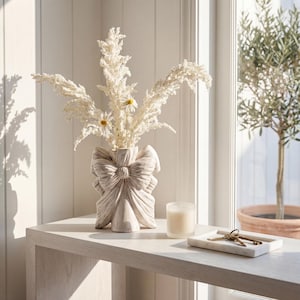 Puede incluir: Un jarrón de cerámica beige con forma de lazo, lleno de flores blancas secas y margaritas. Una vela encendida y una bandeja de mármol con llaves están sobre una mesa de madera clara. La escena está iluminada por luz natural.
