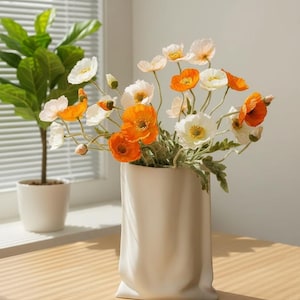 May include: A white ceramic vase filled with orange and white poppy flowers. The vase has a unique, folded design. A potted plant is visible in the background, near a window with blinds. The flowers add a pop of color to the room.