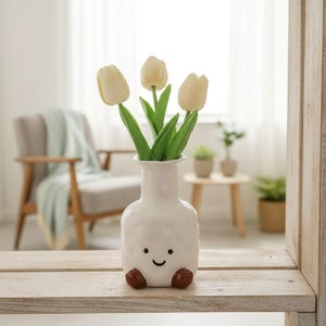 May include: A white ceramic vase with a smiling face and brown feet holds three cream-colored tulip flowers with green stems and leaves. The vase sits on a wooden surface, with a blurred background of a chair and a window.