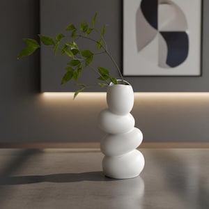 Może przedstawiać: Biały ceramiczny wazon o zaokrąglonym, warstwowym kształcie, z zielonymi, liściastymi gałązkami. Wazon ma gładkie, błyszczące wykończenie i stoi na neutralnej powierzchni. Wazon ma około 25 cm wysokości.