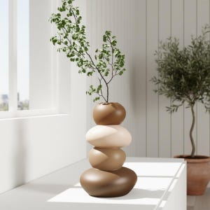 Puede incluir: Un jarrón de cerámica apilado en tonos marrones y beige, que se asemeja a una torre de piedras. El jarrón contiene una ramita de vegetación. El jarrón está sobre una superficie blanca, con una ventana y un pequeño árbol en maceta en el fondo.
