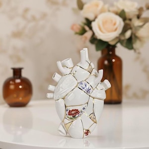 May include: A white ceramic anatomical heart sculpture featuring a kintsugi-style design with gold-colored seams connecting various porcelain fragments. Some segments display delicate floral patterns in red, blue, and green hues on the surface.