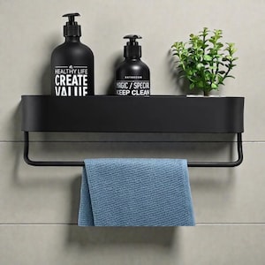 Könnte beinhalten: Schwarzes Badezimmerregal mit Handtuchhalter, mit zwei schwarzen Pumpflaschen und einer kleinen grünen Pflanze. Die Flaschen tragen weißen Text: "HEALTHY LIFE CREATE VALUE" und "BATHROOM MAGIC / SPECIAL KEEP CLEAN". Ein blaues Handtuch hängt darunter.